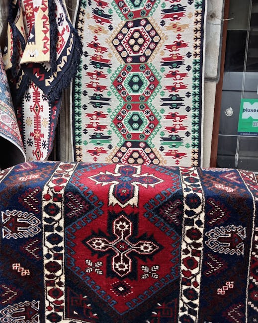 The image shows a display of intricately patterned, traditional woven rugs with geometric designs in rich colors such as red, navy blue, black, and white. The rugs are hanging vertically and draped ov