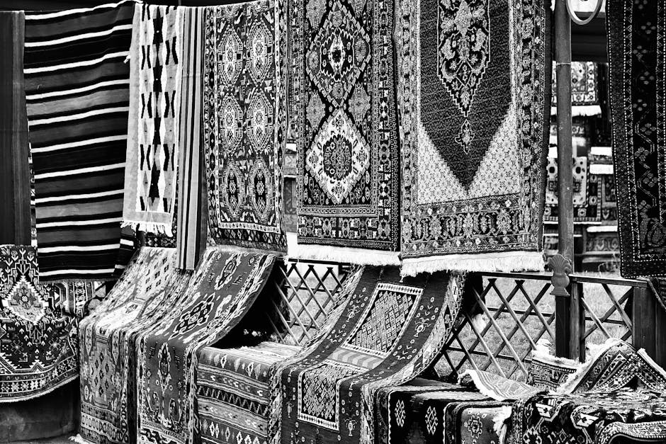 The image shows a display of intricately patterned, traditional woven rugs with geometric designs in rich colors such as red, navy blue, black, and white. The rugs are hanging vertically and draped ov