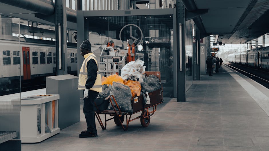 A person in a high-visibility safety vest, dark clothing, and a beanie hat stands on a train station platform beside a flatbed trolley filled with garbage bags and discarded materials, including plast